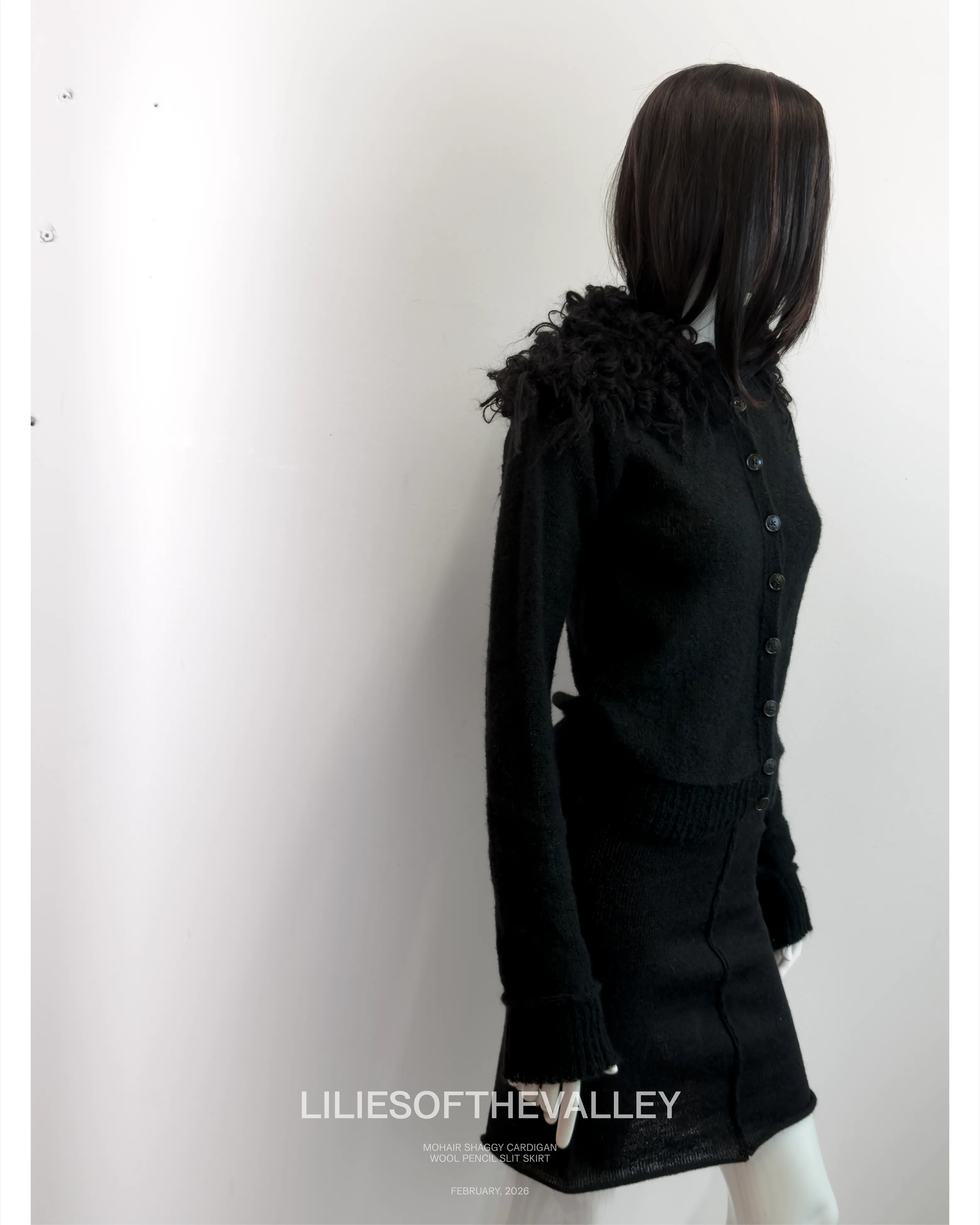 Campaign frame: a black knitted cardigan with a ruffled shoulder, shot
                    side on at full length.