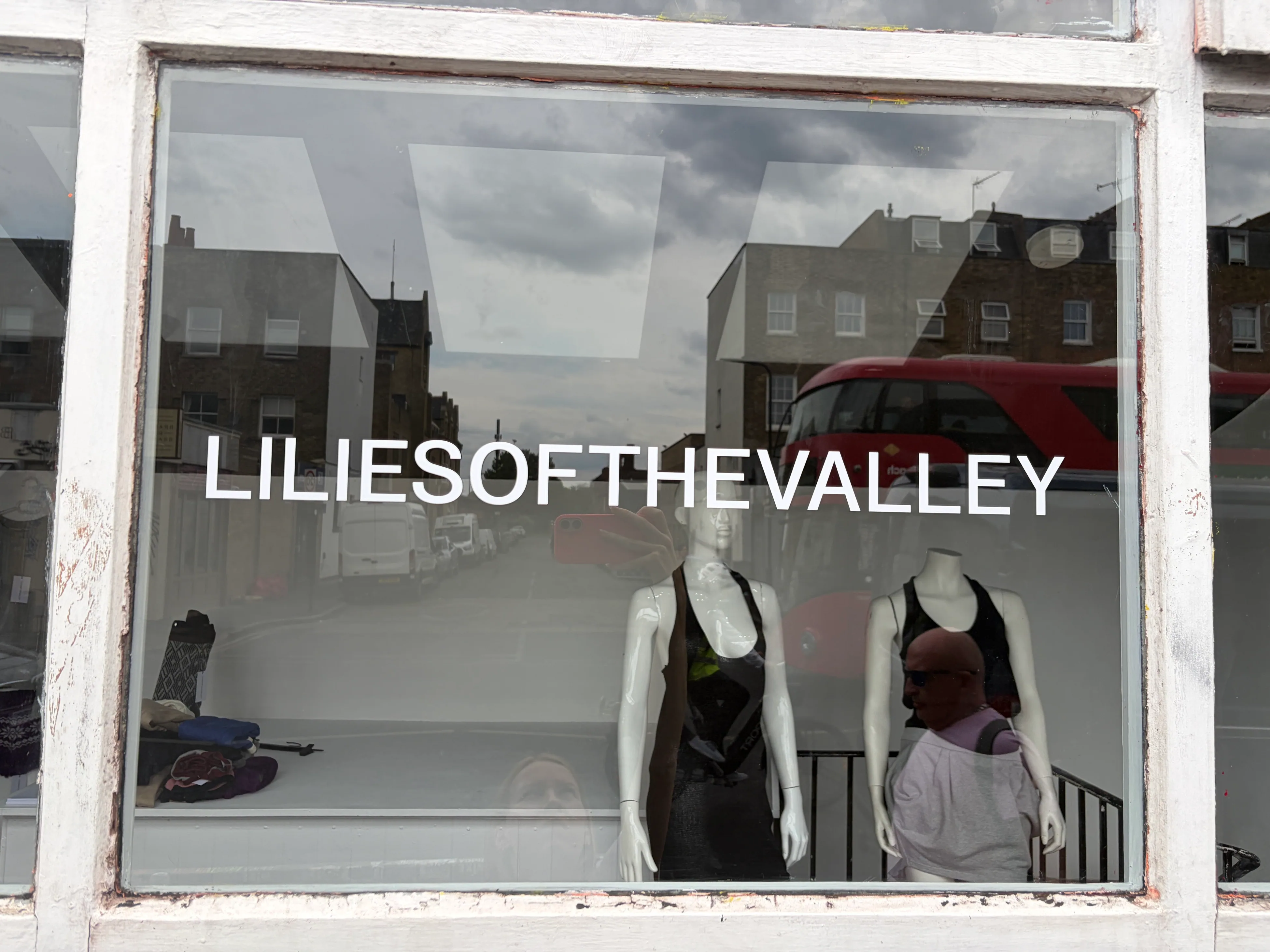 The shopfront at WASTE! SPACE: LILIESOFTHEVALLEY in white vinyl across
                    the glass, two mannequins behind it, a London bus reflected in the pane.