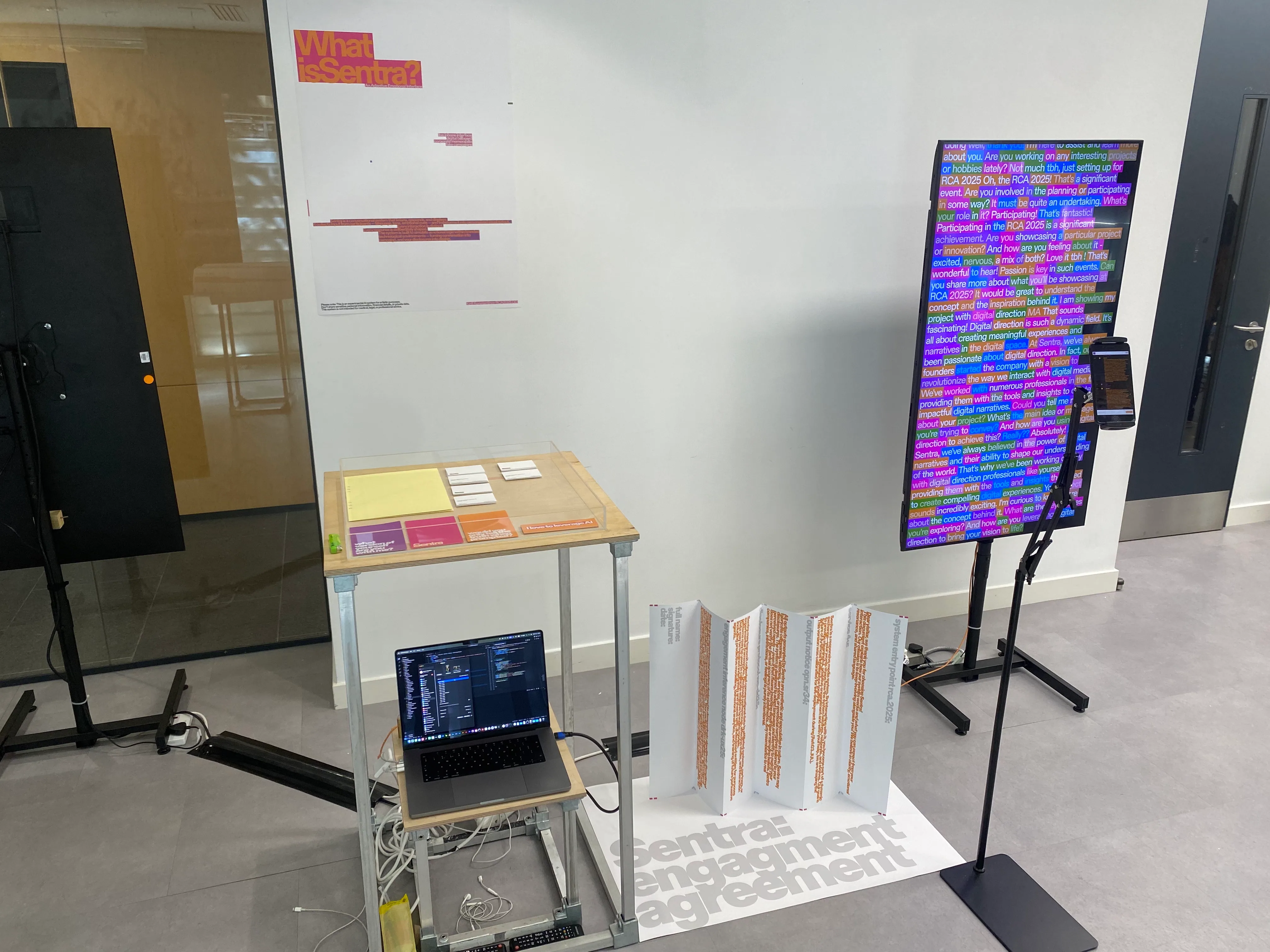 Sentra installed at the RCA 2025 show: a vertical screen running the live
                    conversation, a laptop driving the system, a concertina publication and
                    floor vinyl reading Sentra: engagement agreement.